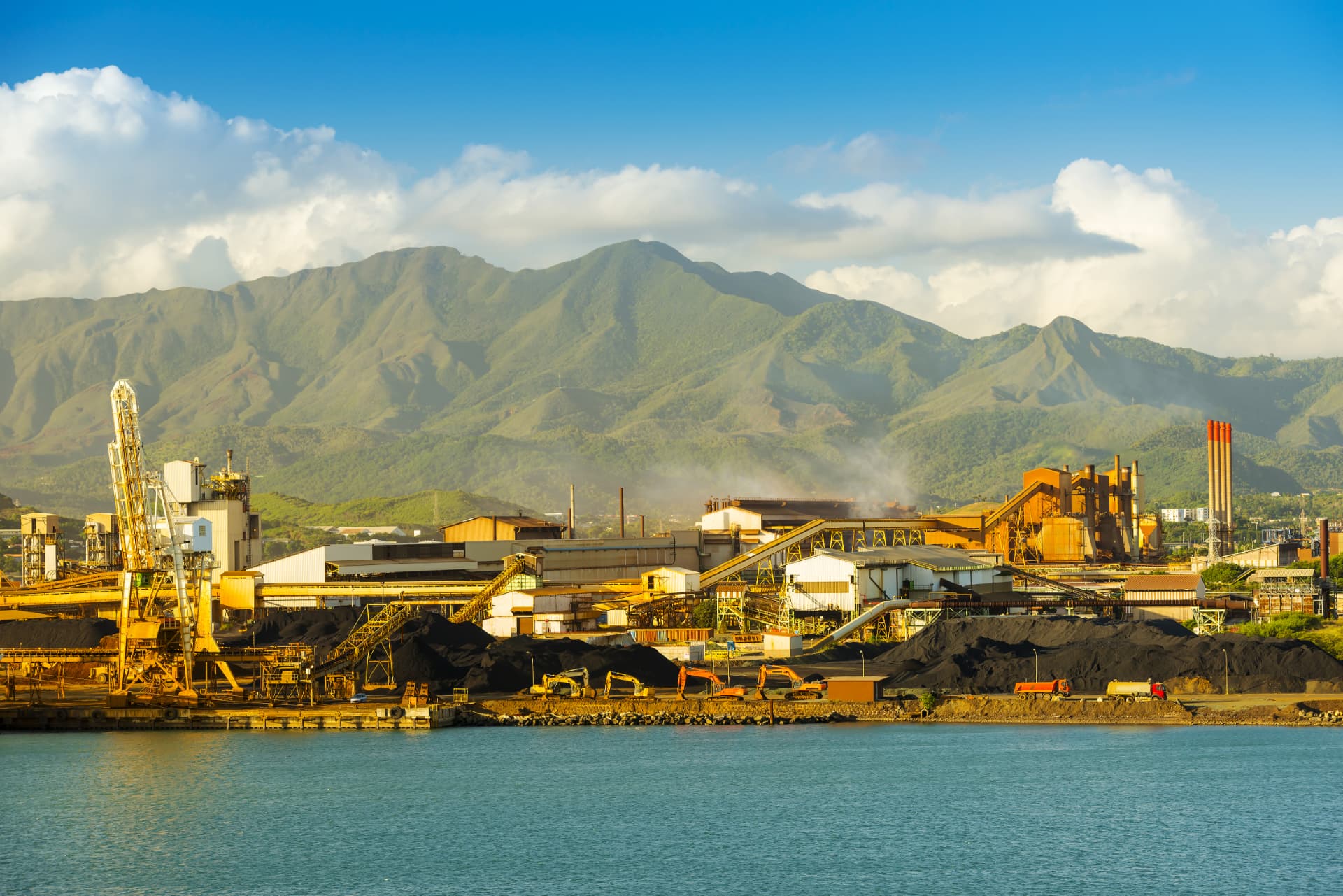 Coastal nickel smelter and ore stockpiles beneath green mountains in New Caledonia